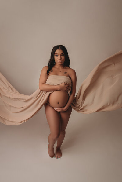 Very pregnant woman posing in an Atlanta maternity portrait studio wearing nude chiffon fabric flowing off of her while she shows her baby bump. Shot on gray backdrop