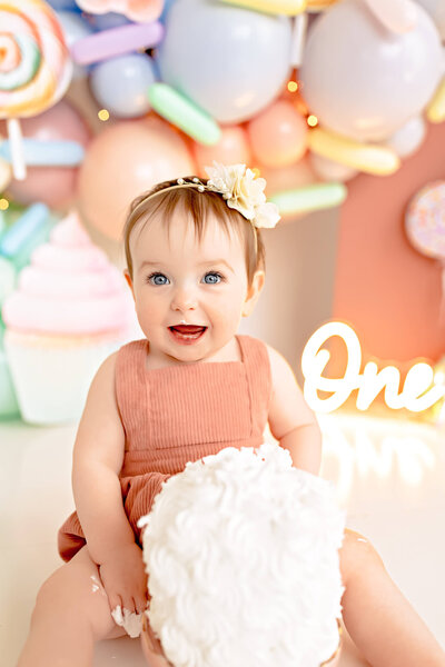 Toddler girl celebrating her first birthday with a cake smash. She is sitting in front of a colorful balloon backdrop.