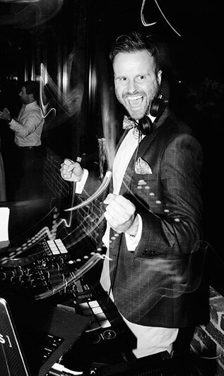 Energetic male DJ in a bow tie and suit playing music behind the decks at an event, with light trails adding motion.