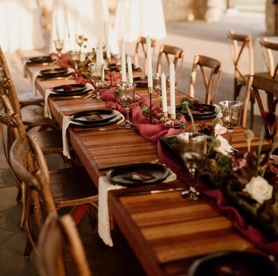 A long wooden table set with black plates, white napkins, long pillar candles, and a red table runner.