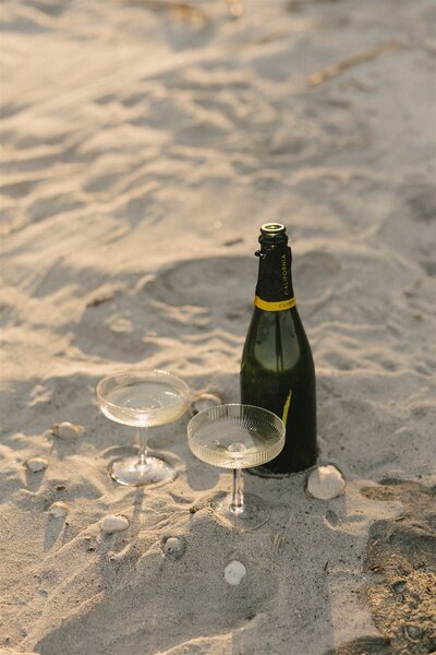A champagne bottle stands in the beach sand with two glasses and shells