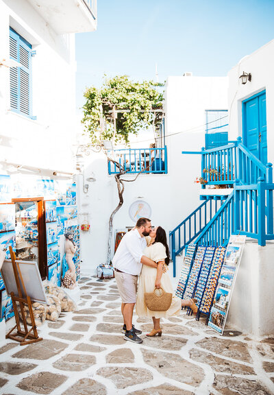 A couple kissing on the cobble stone streets of mykonos with blue stairs and white clay homes around them.