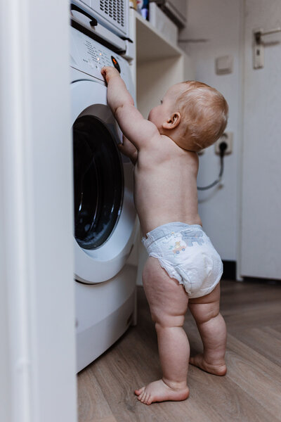 Foto van zijkant baby staat in pamper voor wasmachine op knopjes te drukken