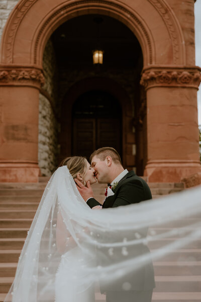 A couple kisses passionately in front of an ornate stone archway. The bride's veil flows gracefully, capturing a romantic and timeless wedding scene.