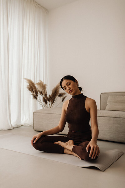  young woman in a deep brown workout set sits cross-legged on a beige yoga mat in her living room, her arms resting on her knees in a meditative pose. Behind her, a neutral-toned sofa and a vase of dried flowers enhance the calming aesthetic. Captured for Elevate Wellness, this image promotes mindfulness, balance, and self-care.