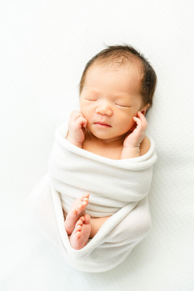 an Asian baby wrapped in a white blanket lays on a white bean bag while asleep during his posed newborn session in Austin. 