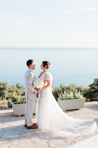 Couple standing together in front of a serene sea view, holding a white bouquet during their romantic coastal wedding
