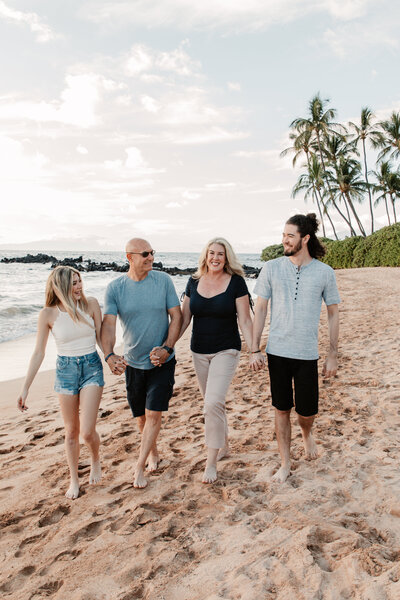 family of four walking along the beach in Maui during a sunset photoshoot holding hands walking along the beach with palm trees in the background