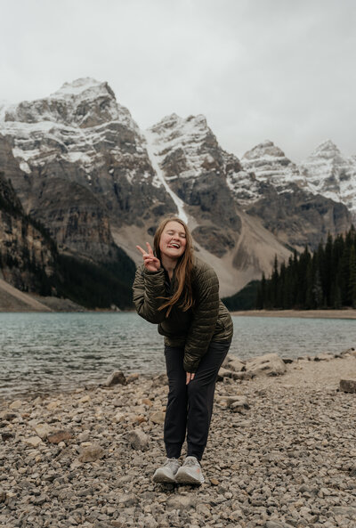 Sydney Breann, a Montana-based elopement photographer, smiles and poses with a peace sign beside Moraine Lake, surrounded by rugged mountain peaks and alpine water.