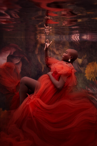 A woman gracefully reclines underwater, adorned in a flowing red tulle gown with ruffled detailing that softly billows around her. Her serene, poised expression complements the luxurious and ethereal atmosphere created by the deep floral backdrop. The vivid red gown contrasts beautifully against the muted tones, enhancing the dreamy underwater scene.