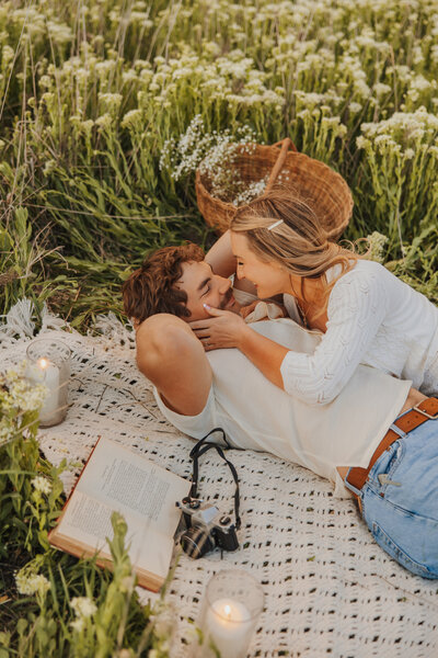 Couple laying on a picnic blanket in a field of white flowers, smiling at each other during an outdoor session.
