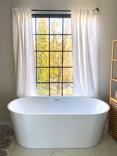 Freestanding soaking tub in a bright bathroom with large window and white curtains.