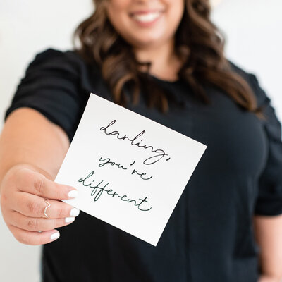 In this portrait, Lena, the owner of Lena Designs, holds a card with the quote "Darling, you're different" written on it in cursive style.