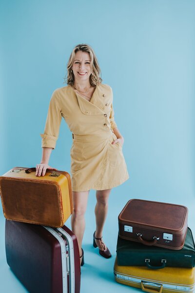 Woman with shoulder-length blonde stands near several vintage suitcases and smiles at the camera in front of an aqua backdrop.