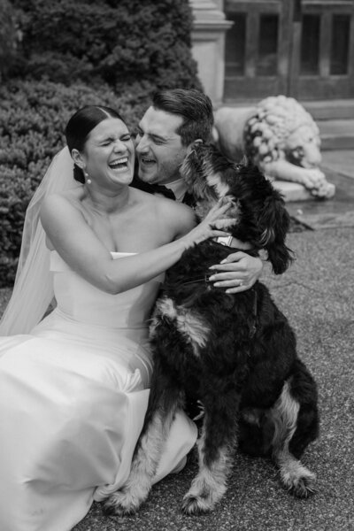 A newlywed couple sitting on the ground laughing with their dog 