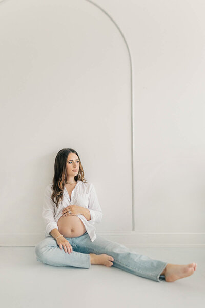 Maternity portrait of a pregnant woman sitting on the floor in a white button-down shirt and jeans, gently holding her baby bump in a Kansas City studio.