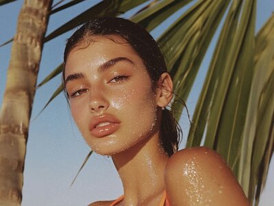 Close-up of a woman with wet hair and dewy skin, set against palm leaves and a blue sky, exuding a fresh, tropical vibe.