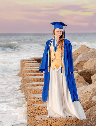 A Cape Fear Communicy College graduating senior in a blue cap and gown standing confidently on beach rocks with waves crashing in Wilmington, North Carolina. This striking image captures the excitement of graduation, ideal for artistic senior photography by the ocean.