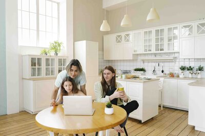 A family of three sit around a kitchen table, with whiete kitchen cabinets and houseplants in the background. The two parents are androgynous with long hair, and both are wearing pastel colored shirts. They are smiling at a laptop screen, being shown to them by their child, who sits in the middle of the table.