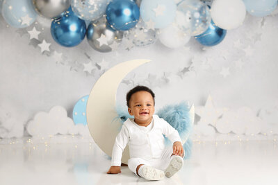 A happy baby boy sits on a crescent moon prop, dressed in a white outfit, surrounded by blue and silver star-themed decorations.