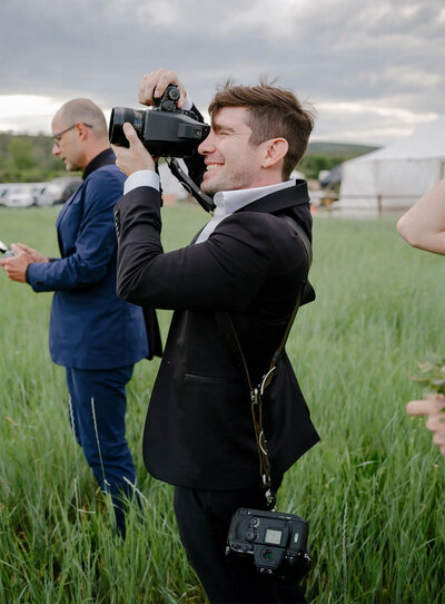 Lance in a black suit takes a photo with a camera in a grassy field. Another man in a blue suit stands nearby holding a phone.