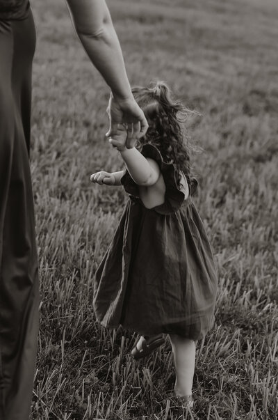 A black and white photo captures a New England family moment: a young child with curly hair in a ruffled dress, holding an adult's hand, walking on grass. This portrait, reminiscent of documentary-style photography across New England, beautifully frames the guiding bond between generations.