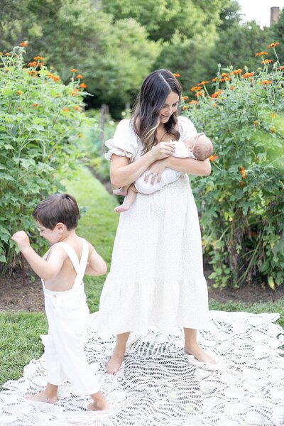 Mom Standing in a garden surrounded by flowers holding her newborn while her 3 year old son runs past, Virginia Beach Family Photography