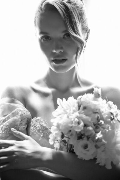 A dramatic black-and-white portrait of a bride holding a white floral bouquet, wearing an off-the-shoulder lace gown, captured in soft natural light
