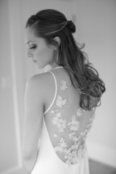 Black and white photo of bride looking over her shoulder, showcasing intricate back detail of her wedding dress. Deborah Zoe Photography