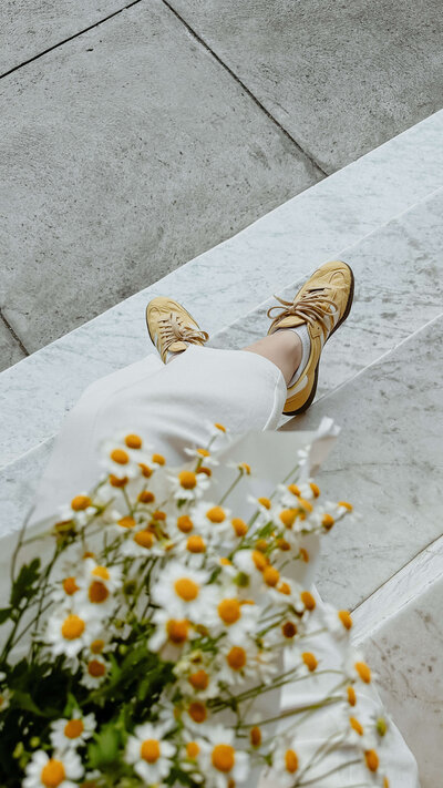 Person sitting on stone steps wearing yellow sneakers and white pants, holding a bouquet of daisies, looks deep in thought as if contemplating the next chapter of their memoir.