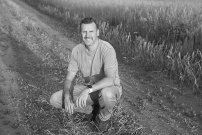 Lon Stroschein kneeling on a dirt path in a field, smiling at the camera