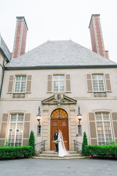 A bride and groom stand on the steps outside the front of Glen Manor House. 