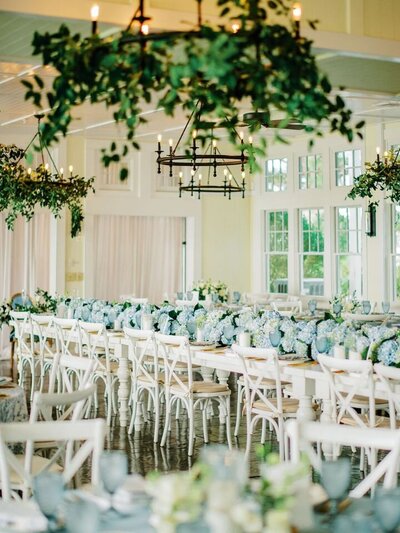 A reception hall filled with blue hydrangeas, white farm tables and white crossback chairs