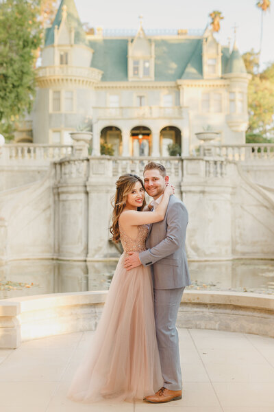 Kohnur Bride and groom dancing in front of Kimberly Crest House and Gardens in Redlands, California during a whimsical castle-style engagement session featuring soft glam bridal makeup and half-up loose waves hairstyle by Kohnur