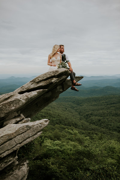 bride and groom sitting on cliff with mountains in background