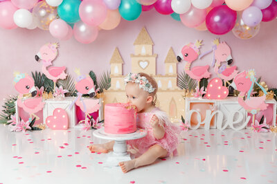 Little girl in a pink feathery outfit biting into a pink cake surrounded by a castle, balloons and princess flamingos.
