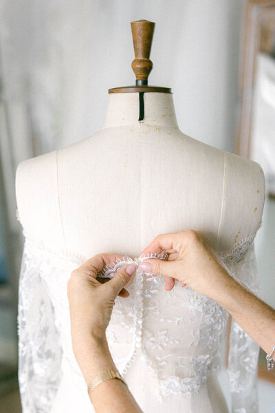 Bridal seamstress fastening delicate lace buttons on a handmade wedding gown displayed on a dress form.