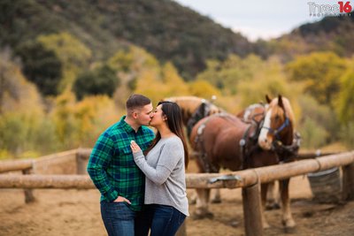 Engaged couple share a kiss during engagement session at Oak Glen Preserve while a horse looks on