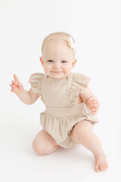 One year old baby in beige romper with neutral colored bow on head is smiling at the camera and holding up one finger at her first birthday photo session in portland, oregon