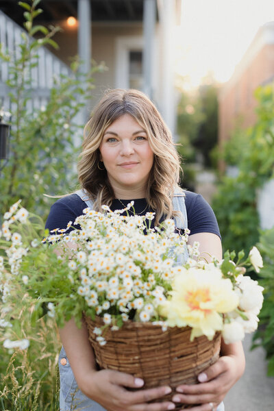 Light skinned blonde woman carrying a large basket of yellow and white flowers with both hands. Photo is close up and she is looking at camera and smiling, she is standing in a garden