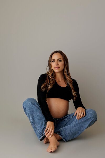 A pregnant woman sits on the floor with her legs crossed, wearing a black long-sleeve crop top and loose-fitting blue jeans. Her baby bump is visible, and she has long, wavy hair cascading over her shoulders. She gazes calmly at the camera with a serene expression. The background is a soft, neutral gray, adding to the simplicity and elegance of the portrait.