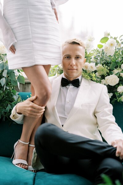 Groom in white tuxedo sitting on a green velvet couch, confidently holding the bride's leg as she stands beside him in a chic short wedding dress, surrounded by white roses and greenery