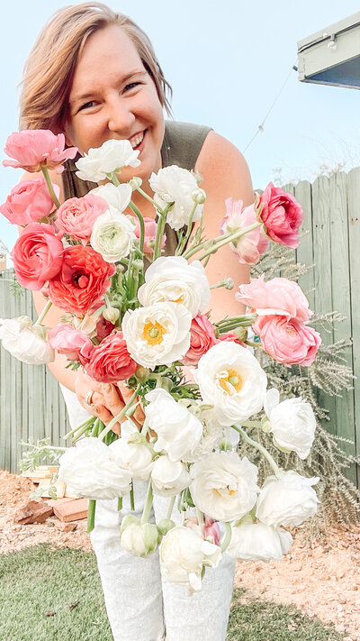Photo of Sam Lynn Photo holding a bunch of pink and white ranunculus leaning in and smiling at the camera