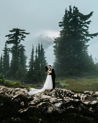 a couple in wedding attire embrace on top of a boulder. Deep, green pines tower behind them and through the dense fog, we see the peaks of the cascade range peek out.  