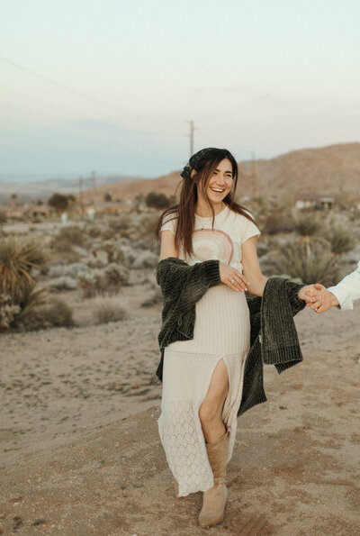 A joyful woman in a white dress and green cardigan holds hands with someone off-camera, smiling and walking through a desert landscape at dusk.