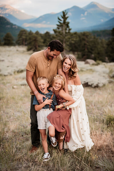 A family of four laughing together in Rock Mountain National Park field while the sun sets behind the mountains. 