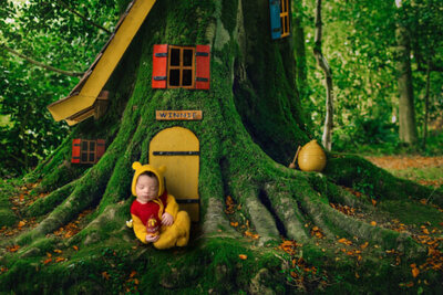 Newborn boy dressed as Pooh Bear sleeps beside a tree
