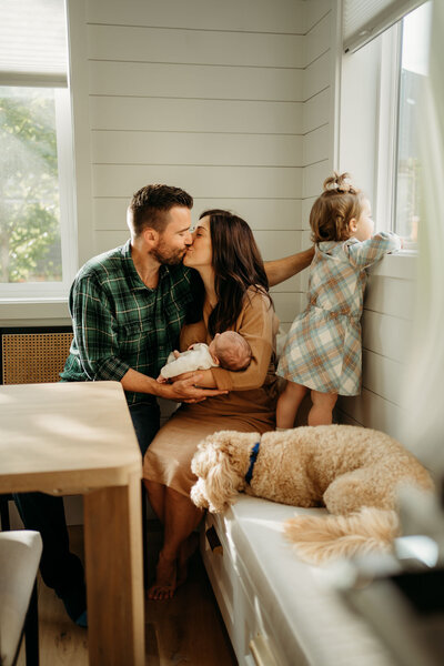 Dad and mom kissing while they hold their newborn together at their dinning  table