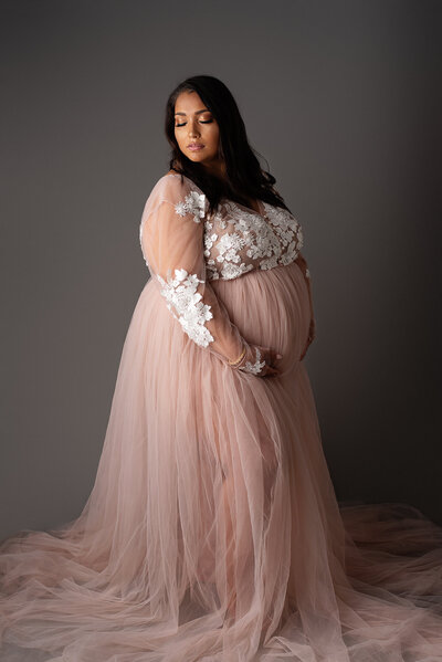 Studio maternity portrait of mom in pink dress holding belly against gray backdrop in Jacksonville, FL.