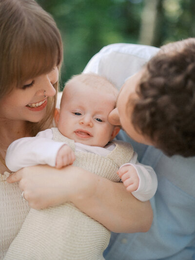 Mother and father kissing and playing with their newborn baby at a park in Squamish